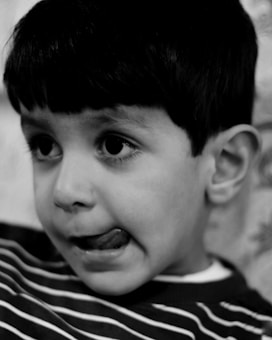 A close-up black and white portrait of a young child with short dark hair and expressive eyes. The child is wearing a striped shirt and has their tongue slightly out, enhancing an innocent and playful expression.