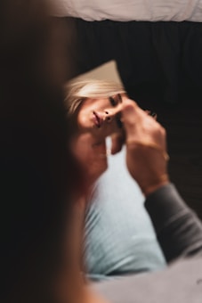 A woman's face is partially reflected in a small, handheld mirror. She appears to be in a casual setting, holding the mirror in one hand. The focus is on her face, while the background is blurred. The lighting is soft, creating a serene and intimate atmosphere.