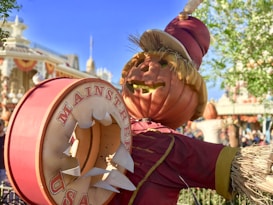 A pumpkin-headed scarecrow is dressed in a red costume with straw accents and is holding a large round sign with the text 'Main Street USA' on it. The background shows a festive setting with a building decorated with red, orange, and yellow banners, and a clear blue sky with some trees.