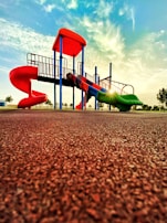 A vibrant playground with a slide and climbing structures, featuring vivid red and green colors, is set against a backdrop of a bright sky with scattered clouds. The sun casts a warm glow, creating a serene and inviting atmosphere.