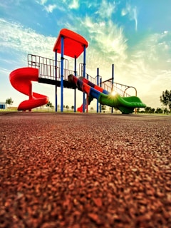 A vibrant playground with a slide and climbing structures, featuring vivid red and green colors, is set against a backdrop of a bright sky with scattered clouds. The sun casts a warm glow, creating a serene and inviting atmosphere.