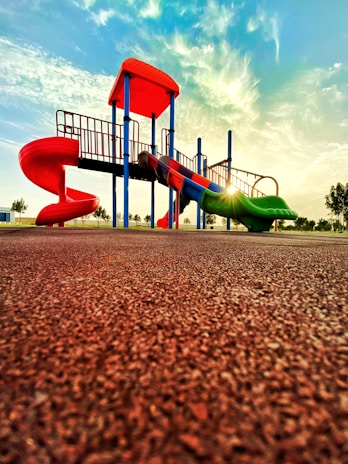 A vibrant playground with a slide and climbing structures, featuring vivid red and green colors, is set against a backdrop of a bright sky with scattered clouds. The sun casts a warm glow, creating a serene and inviting atmosphere.