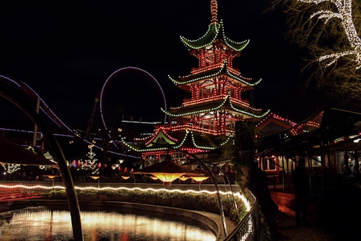 A brightly lit, multi-tiered pagoda stands as the centerpiece, adorned with colorful lights outlining its edges, against a nighttime sky. Surrounding the pagoda are additional light displays, including festive string lights on trees and paths. In the foreground, a curved line of reflective water is bordered by illuminated fencing, creating a vibrant and lively atmosphere.