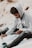 A person sits outdoors holding a paintbrush in one hand and a phone in the other, appearing focused on painting. The individual wears a gray hoodie and dark jeans and is surrounded by art supplies, including a jar of paint.