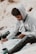 A person sits outdoors holding a paintbrush in one hand and a phone in the other, appearing focused on painting. The individual wears a gray hoodie and dark jeans and is surrounded by art supplies, including a jar of paint.