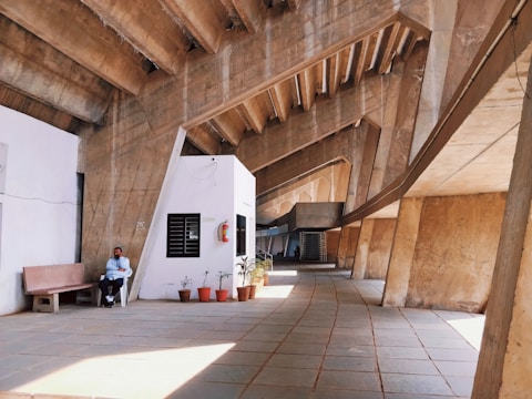 A modern architectural space featuring large concrete beams and an open hallway. A person is seated on a bench near a wall with potted plants and a fire extinguisher. The floor is tiled and the space has a geometric, angular design.