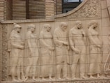 The image depicts a classical relief sculpture featuring a group of six figures depicted in a naturalistic style. They are dressed in ancient garments, and their postures convey a sense of calm and reflection. Intricate architectural details, including patterns and floral motifs, are present in the background.