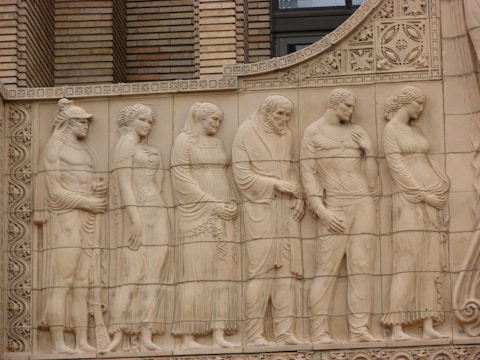 The image depicts a classical relief sculpture featuring a group of six figures depicted in a naturalistic style. They are dressed in ancient garments, and their postures convey a sense of calm and reflection. Intricate architectural details, including patterns and floral motifs, are present in the background.