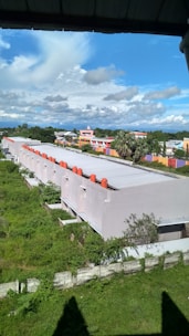 A row of uniform white buildings with red structures on the roof, situated in a green grassy area. Behind the buildings, a colorful residential area features bright orange and purple walls. A clear blue sky with scattered white clouds forms the backdrop.