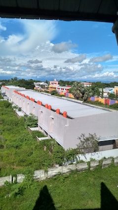 A row of uniform white buildings with red structures on the roof, situated in a green grassy area. Behind the buildings, a colorful residential area features bright orange and purple walls. A clear blue sky with scattered white clouds forms the backdrop.