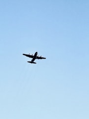 A large propeller aircraft flying in the clear blue sky with minimal clouds. The aircraft is silhouetted against the sky, highlighting its wings and propellers.