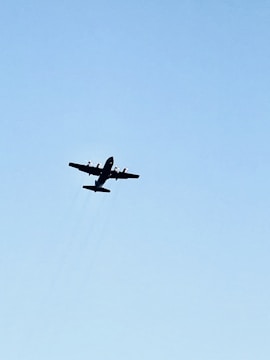 A large propeller aircraft flying in the clear blue sky with minimal clouds. The aircraft is silhouetted against the sky, highlighting its wings and propellers.
