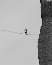 A person is balancing on a tightrope suspended between a rocky cliff and another point, high above the ground. The scene is monochrome, emphasizing the contrast between the person's silhouette and the rugged texture of the rock face.