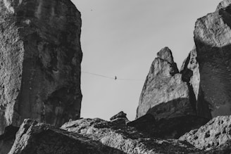 A person is walking on a tightrope stretched between two towering rock formations. The scene conveys a sense of daring and skill against a stark backdrop. The rugged texture of the rocks is accentuated by shadows and light, creating a dramatic effect.