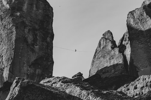 A person is walking on a tightrope stretched between two towering rock formations. The scene conveys a sense of daring and skill against a stark backdrop. The rugged texture of the rocks is accentuated by shadows and light, creating a dramatic effect.