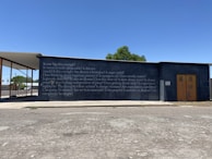A concrete wall with a series of philosophical questions written on it in white text, set against a clear blue sky. A structure extends from the left side, partially covering the wall. There is a small tree visible in the background and a road or parking area with a patchy asphalt surface in the foreground.