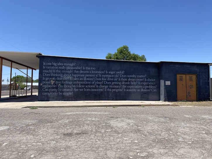 A concrete wall with a series of philosophical questions written on it in white text, set against a clear blue sky. A structure extends from the left side, partially covering the wall. There is a small tree visible in the background and a road or parking area with a patchy asphalt surface in the foreground.