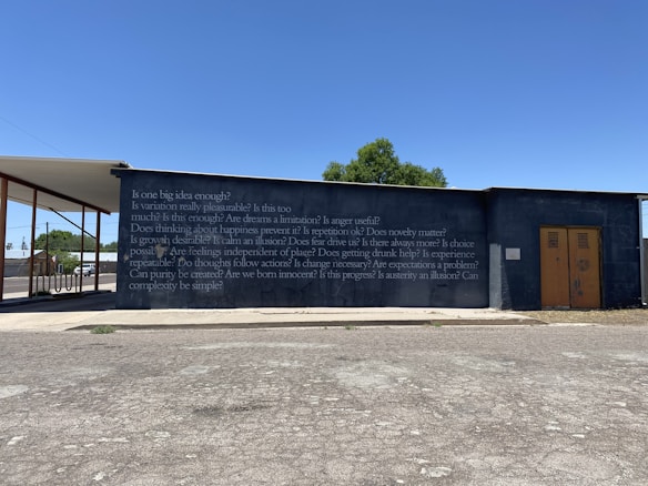A concrete wall with a series of philosophical questions written on it in white text, set against a clear blue sky. A structure extends from the left side, partially covering the wall. There is a small tree visible in the background and a road or parking area with a patchy asphalt surface in the foreground.