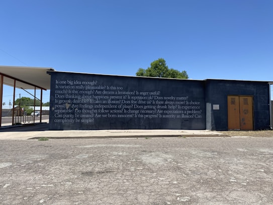 A concrete wall with a series of philosophical questions written on it in white text, set against a clear blue sky. A structure extends from the left side, partially covering the wall. There is a small tree visible in the background and a road or parking area with a patchy asphalt surface in the foreground.