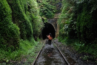 A person with a backpack walks along an abandoned railway track surrounded by lush green vegetation leading to a dark, arched tunnel entrance. The scene is enveloped in dense greenery, creating a sense of mystery and wilderness.