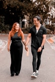 A smiling couple walking hand in hand along a road, surrounded by greenery. Both are dressed in black, complementing each other's attire. Their expressions suggest joy and comfort in each other's company.