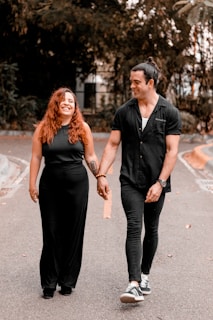 A smiling couple walking hand in hand along a road, surrounded by greenery. Both are dressed in black, complementing each other's attire. Their expressions suggest joy and comfort in each other's company.