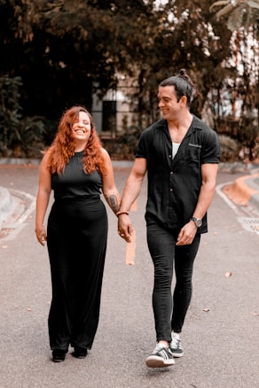 A smiling couple walking hand in hand along a road, surrounded by greenery. Both are dressed in black, complementing each other's attire. Their expressions suggest joy and comfort in each other's company.