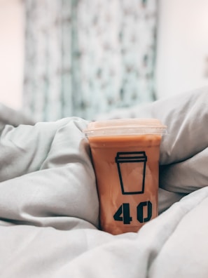 A plastic cup with an iced beverage rests on a bed covered with light-colored blankets. The cup features a design of a coffee cup and the number 40 printed in black. The background is blurred, suggesting curtains or a window with some light coming through.