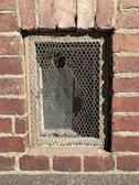 A small rectangular window set in a red brick wall is covered by a mesh wire screen, possibly for protection or ventilation. The bricks are weathered with visible mortar lines, and sunlight casts shadows, highlighting the rough texture of both the bricks and the wire mesh.