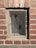 A small rectangular window set in a red brick wall is covered by a mesh wire screen, possibly for protection or ventilation. The bricks are weathered with visible mortar lines, and sunlight casts shadows, highlighting the rough texture of both the bricks and the wire mesh.