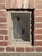 A small rectangular window set in a red brick wall is covered by a mesh wire screen, possibly for protection or ventilation. The bricks are weathered with visible mortar lines, and sunlight casts shadows, highlighting the rough texture of both the bricks and the wire mesh.