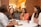 Four women are gathered around a table in a cozy café setting, engaged in conversation and smiling. The space features brick walls and warm lighting creating an inviting atmosphere. Each person has a beverage in front of them, signaling a casual and friendly meeting.