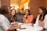 Four women are gathered around a table in a cozy café setting, engaged in conversation and smiling. The space features brick walls and warm lighting creating an inviting atmosphere. Each person has a beverage in front of them, signaling a casual and friendly meeting.