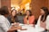 Four women are gathered around a table in a cozy café setting, engaged in conversation and smiling. The space features brick walls and warm lighting creating an inviting atmosphere. Each person has a beverage in front of them, signaling a casual and friendly meeting.