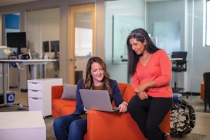 Two women are engaged in a discussion while looking at a laptop screen in a modern office setting. One woman is sitting on an orange couch, and the other is standing beside her, leaning slightly on the couch. The office environment includes a standing desk with monitors and chairs.