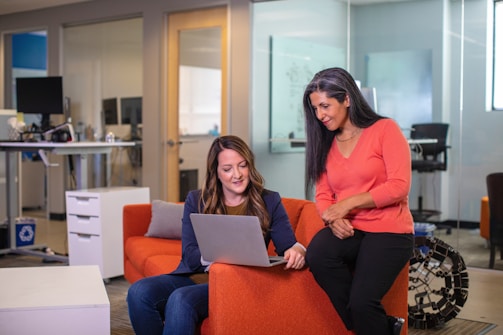 Two women are engaged in a discussion while looking at a laptop screen in a modern office setting. One woman is sitting on an orange couch, and the other is standing beside her, leaning slightly on the couch. The office environment includes a standing desk with monitors and chairs.