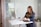 A woman stands in a modern office space by a desk with a large computer monitor and a tablet. She is dressed in a casual business outfit, has long hair, and is looking at her smartphone. The office is bright with some greenery and a glass door visible in the background.