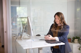 A woman stands in a modern office space by a desk with a large computer monitor and a tablet. She is dressed in a casual business outfit, has long hair, and is looking at her smartphone. The office is bright with some greenery and a glass door visible in the background.