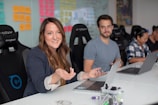 A group of people sitting at a conference table with laptops. The woman in the foreground is gesturing with her hands and smiling, while another person looks on and two others work. The background features a wall with colorful sticky notes.