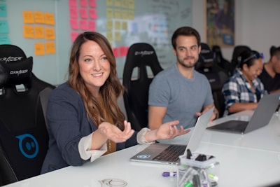 A group of people sitting at a conference table with laptops. The woman in the foreground is gesturing with her hands and smiling, while another person looks on and two others work. The background features a wall with colorful sticky notes.