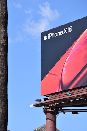 A large billboard for the iPhone XR stands against a clear blue sky. The billboard features a sleek design with the Apple logo and branding in white on a dark background, alongside a vibrant red image of the phone.