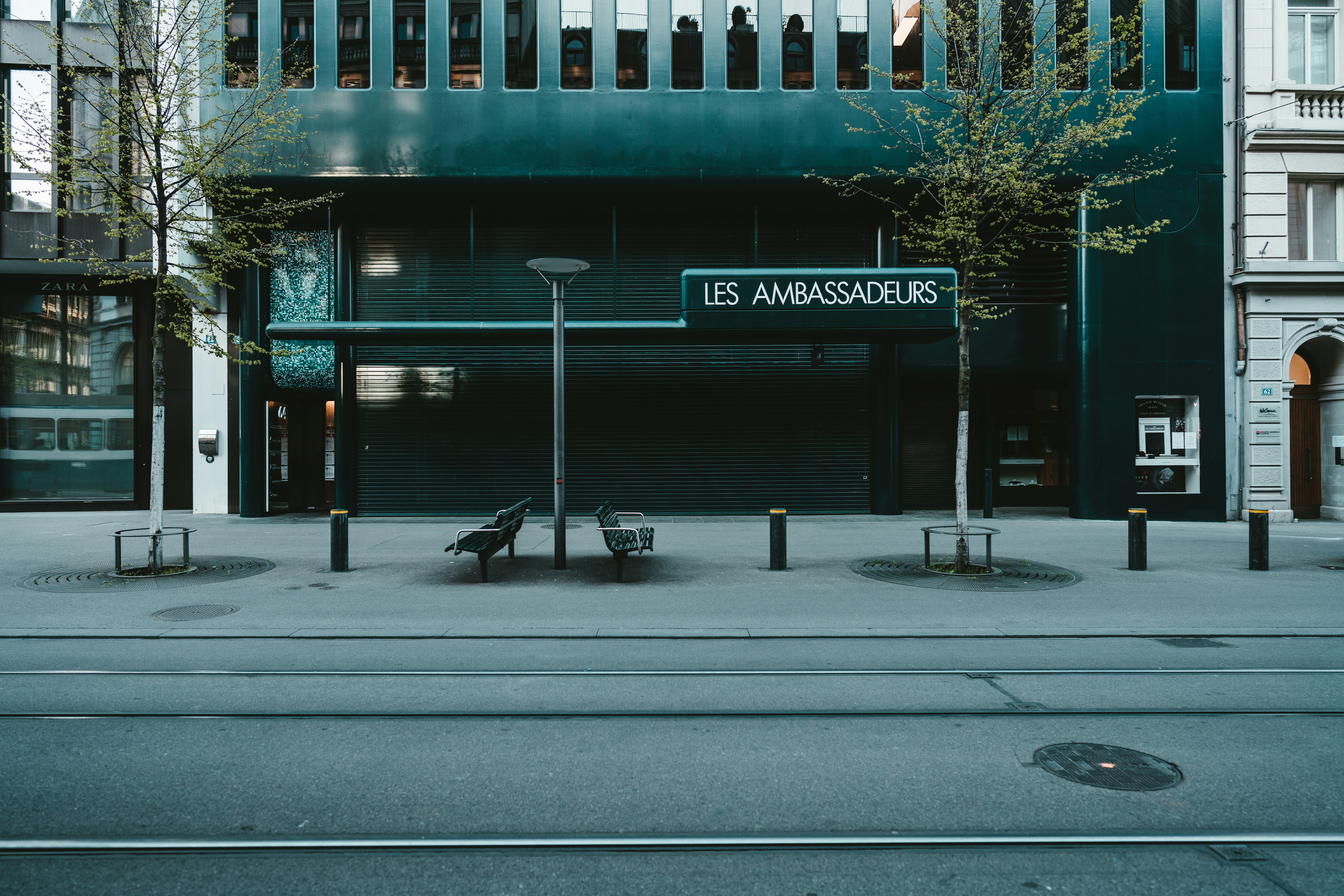 A street scene featuring a modern storefront with the sign 'Les Ambassadeurs.' The building facade is dark with large windows lining the top. Two trees with sparse leaves are evenly spaced in front of the shop. The pavement has designated walking paths, and there are a few benches and lamp posts near the storefront. Tram tracks run parallel to the street.