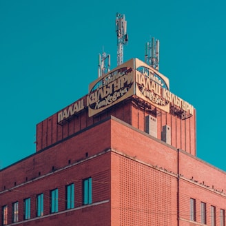 A red brick building with a vintage sign on top displaying Cyrillic letters and surrounded by several metal antennas against a clear blue sky.