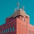 A red brick building with a vintage sign on top displaying Cyrillic letters and surrounded by several metal antennas against a clear blue sky.