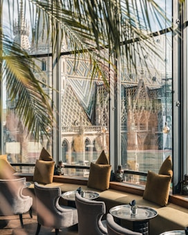A cozy interior with elegant chairs and metallic tables set next to large windows. The windows offer a stunning view of a historic, intricately designed cathedral with a multicolored tiled roof. Palm leaves partially frame the scene, adding a touch of greenery to the sophisticated setting.