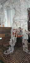 A sculpture made of intricate wire framework depicting a human figure stands on a bed of vertical wooden pegs. Behind the sculpture is a dark wooden chest suggesting a gallery or artistic setting. The wires form curves and shapes that resemble muscles and movement, giving the sculpture a dynamic appearance.