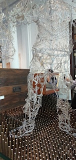 A sculpture made of intricate wire framework depicting a human figure stands on a bed of vertical wooden pegs. Behind the sculpture is a dark wooden chest suggesting a gallery or artistic setting. The wires form curves and shapes that resemble muscles and movement, giving the sculpture a dynamic appearance.