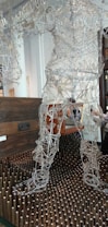 A sculpture made of intricate wire framework depicting a human figure stands on a bed of vertical wooden pegs. Behind the sculpture is a dark wooden chest suggesting a gallery or artistic setting. The wires form curves and shapes that resemble muscles and movement, giving the sculpture a dynamic appearance.