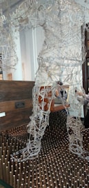 A sculpture made of intricate wire framework depicting a human figure stands on a bed of vertical wooden pegs. Behind the sculpture is a dark wooden chest suggesting a gallery or artistic setting. The wires form curves and shapes that resemble muscles and movement, giving the sculpture a dynamic appearance.