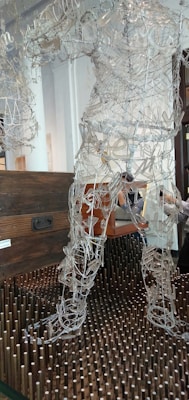 A sculpture made of intricate wire framework depicting a human figure stands on a bed of vertical wooden pegs. Behind the sculpture is a dark wooden chest suggesting a gallery or artistic setting. The wires form curves and shapes that resemble muscles and movement, giving the sculpture a dynamic appearance.